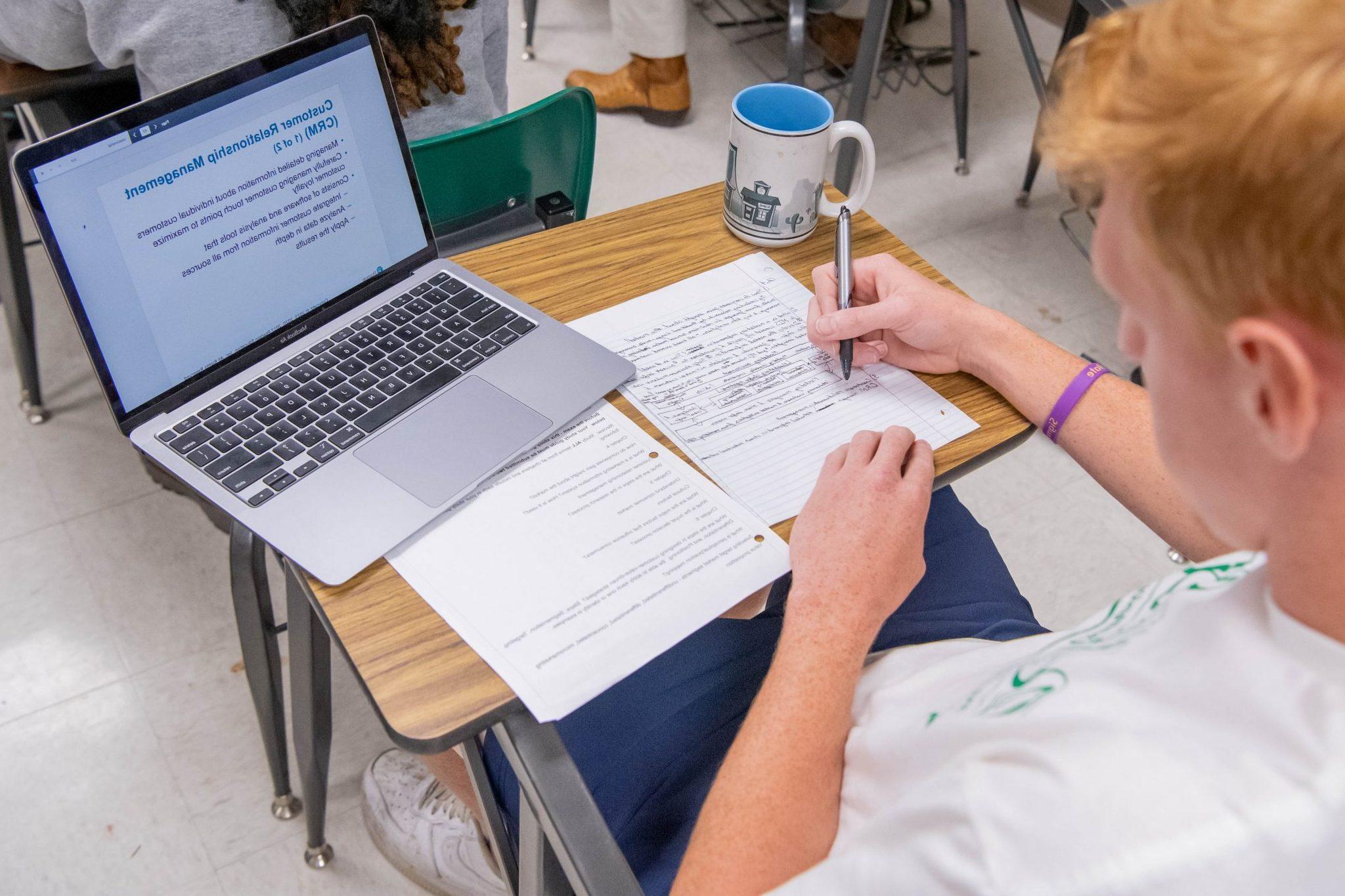 Student sitting at a desk in a classroom, 在纸上做笔记, and looking at a PowerPoint presentation open on his laptop.