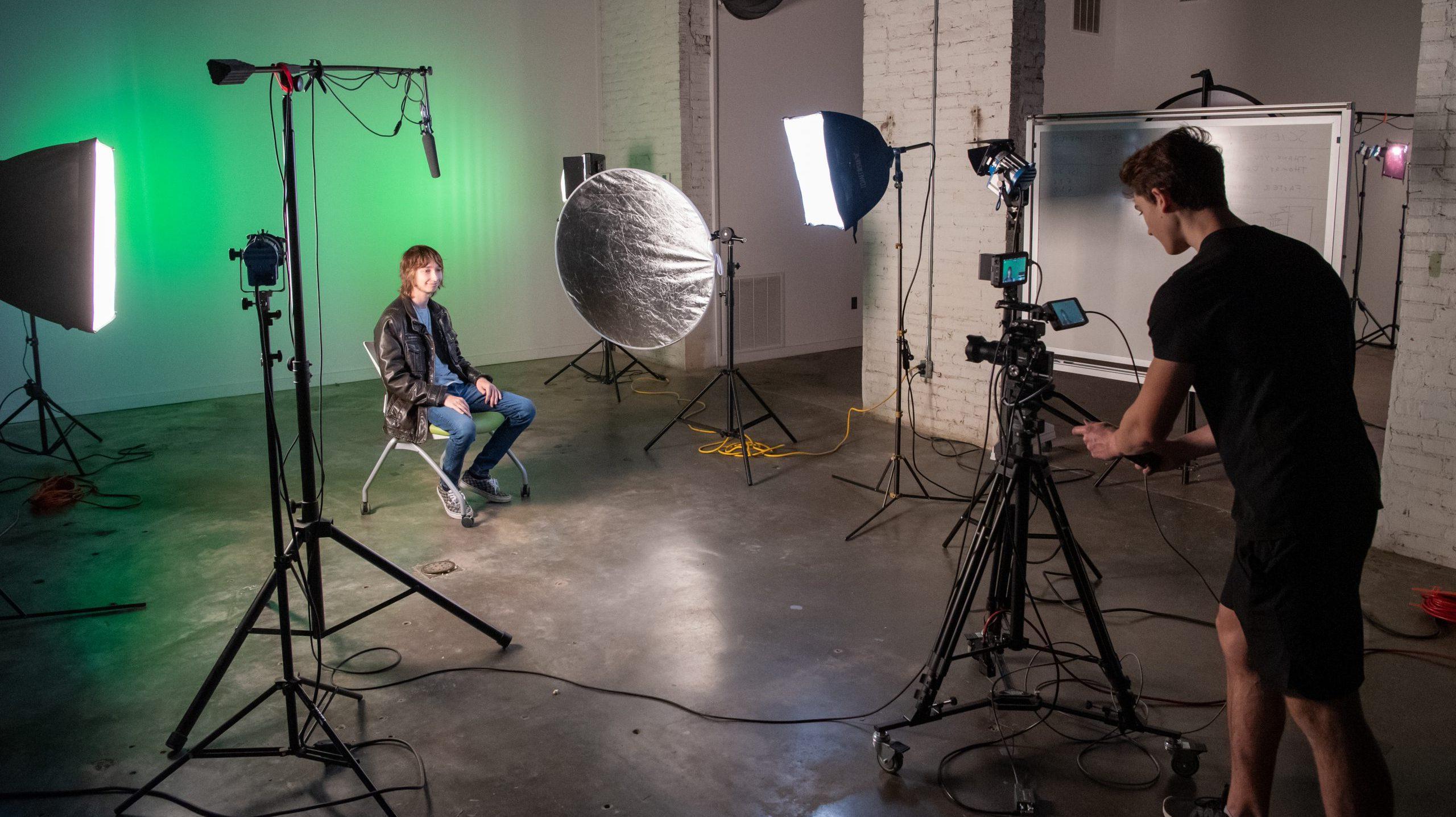 Student sitting in a chair with video equipment and 照明 placed around him, 而另一名学生在三脚架上操作相机.