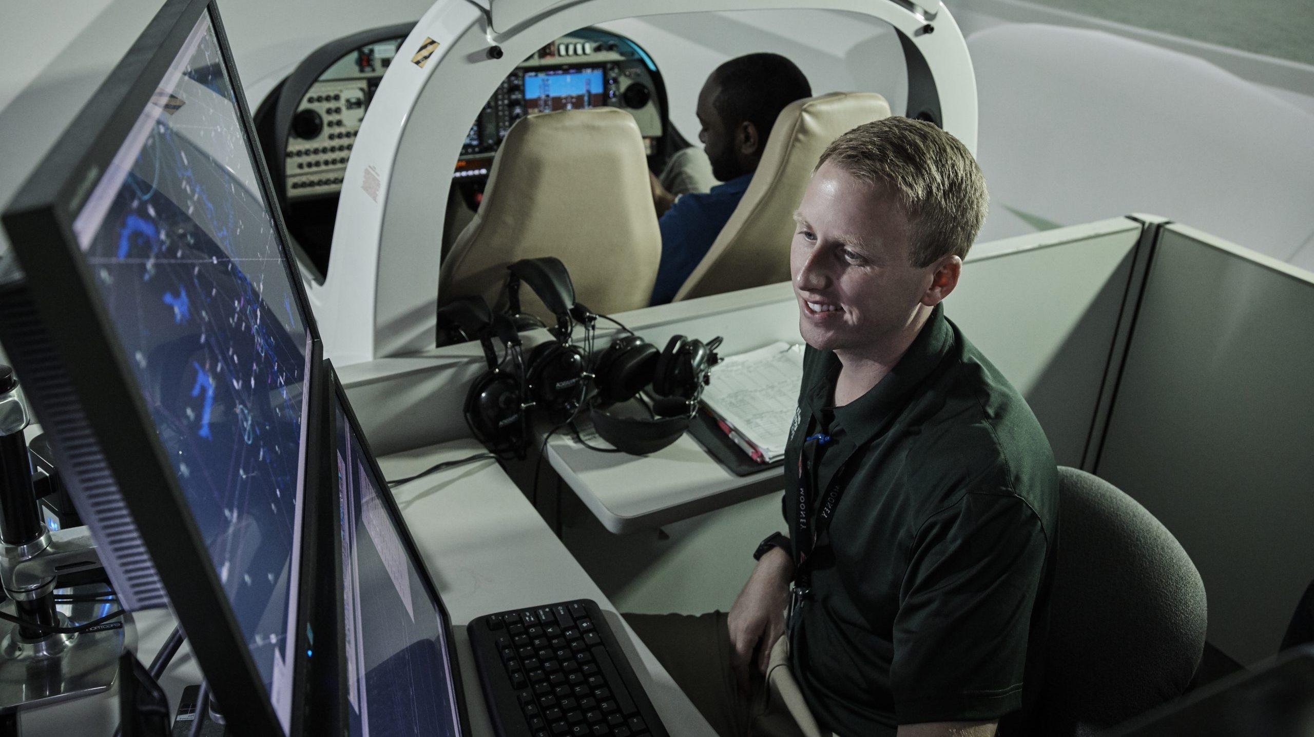 Student looking at computer screens, monitoring air traffic, 而另一个学生在他身后的飞行模拟器里.